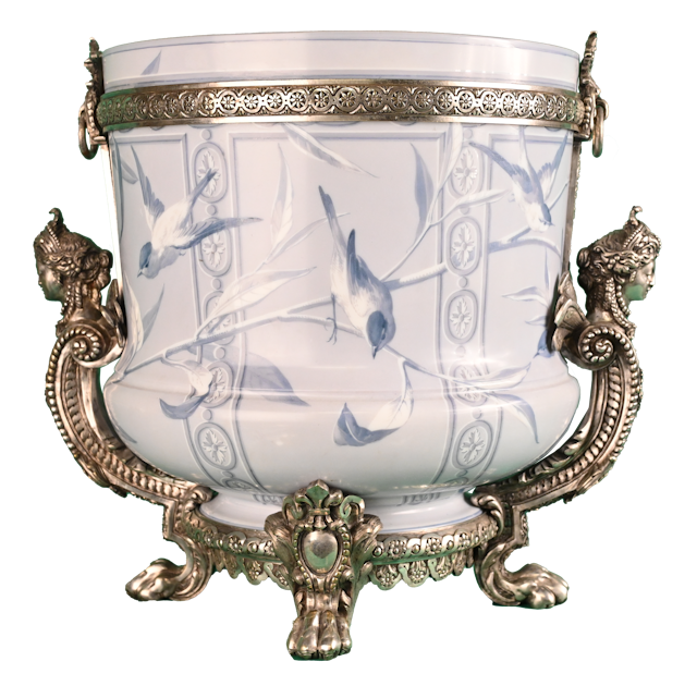 Le vase d’Opaline : la magie de Baccarat au XIXe siècle La cristallerie Baccarat, manufacture de cristal française de renommée internationale, fut fondée sous Louis XV.  Notre vase, réalisé au XIXe siècle, est en opaline. Laiteux, ce verre précieux est orné d’un décor poétique et animalier peint à la main.  Il s’agit de mésanges, symboles du bonheur conjugal et de la fidélité, s’ébattant sur des branches. Ce vase est ceint d’une riche monture en bronze argenté. Elle fut conçue spécifiquement pour cette pièce.  Cet objet richement décoré est le fruit de l’alliance de la magie du verre et du cristal à celle des bronziers spécialisés dans les arts décoratifs. Dits en « pattes de lion », les pieds renvoient à l’imaginaire de la Renaissance, très apprécié au XIXe siècle. La monture est ornée de figures et de détails ornementaux d’un grand raffinement.