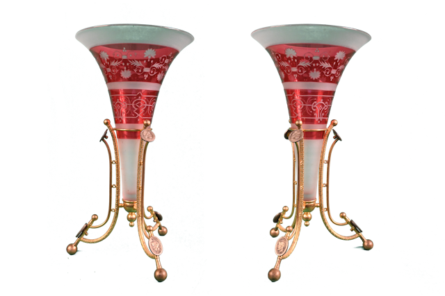 Cette paire de vases, à l’évasement élégant, se caractérise par son alternance colorée.    Celle-ci est obtenue grâce à la création d’un cristal double, transparent vers l’intérieur et rouge vers l’extérieur.    Les bandeaux d’un rouge brillant sont gravés de motifs géométriques et floraux à la roue, tandis que la couche interne, dégagée à l’aide de l’acide ou d’un sablage, à l'aspect mat du verre dépoli.   Ces vases s’inscrivent dans une monture en bronze doré reposant sur trois pieds aux courbes élégantes, achevées par des boules. Des médaillons dans lesquels s’inscrivent des profils de personnages grecs à l’antique sont présents au sommet et à la base de la monture.    Finement ciselée, la monture épouse la forme du vase, et fait écho à la partie haute, en s’évasant vers la base.    Fondée sous Louis XV, la manufacture de cristal française Baccarat fit montre de son excellence aux Expositions Universelles tout au long du XIXe siècle.    La cristallerie Baccarat créa également une coupe sur trépied, dont elle possède encore un exemplaire dans sa collection patrimoniale. S’y côtoient un cristal rouge et un transparent. Comme nos vases, elle est ornée de frises d’inspiration classique et repose sur un trépied en bronze doré. 
